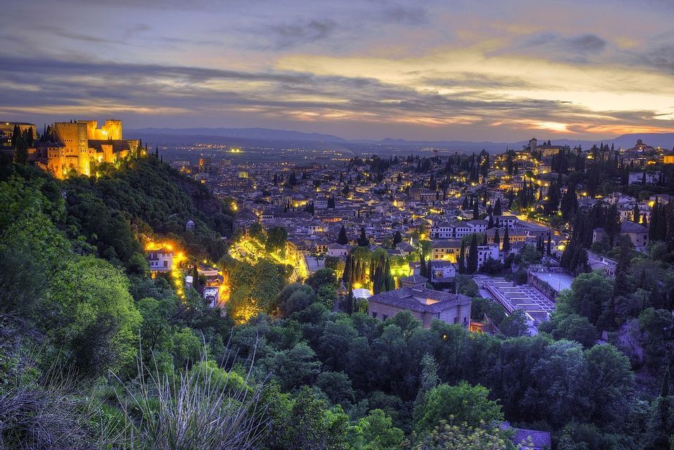 Una fortaleza iluminada en una colina domina un extenso paisaje urbano al anochecer bajo un cielo nublado, con densos árboles verdes en primer plano.
