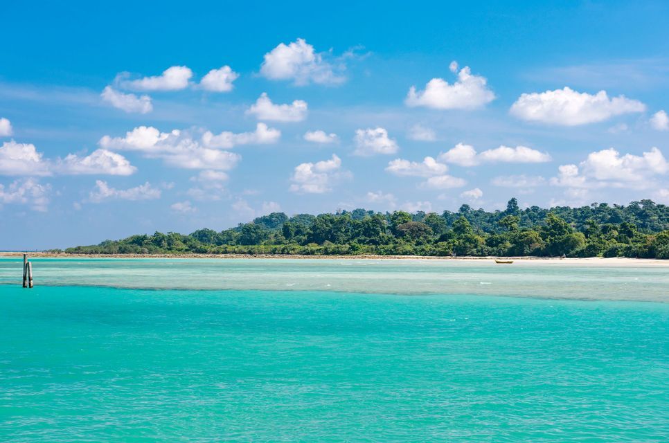 Un'isola rigogliosa e alberata con una spiaggia sabbiosa, visibile attraverso acque limpide e turchesi sotto un cielo azzurro con nuvole bianche.