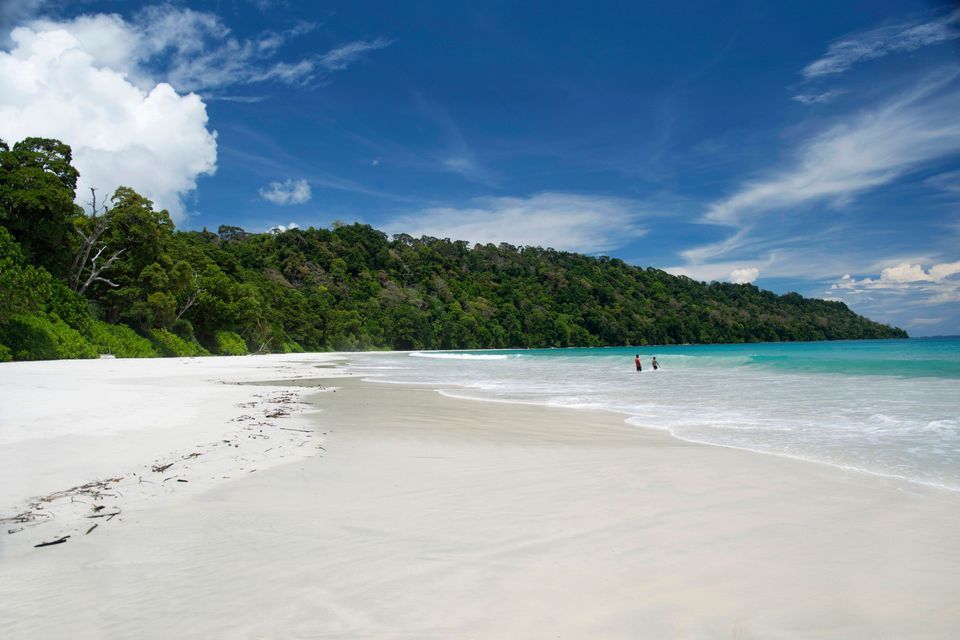 Due persone in piedi in acqua turchese su una spiaggia di sabbia bianca, con una lussureggiante foresta verde sullo sfondo sotto un cielo blu.