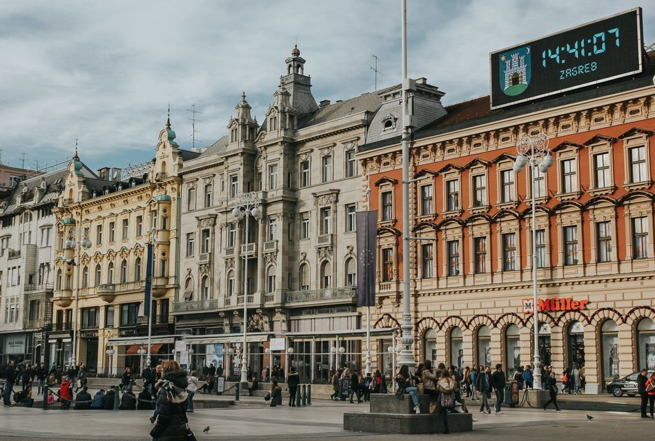 Una vivace piazza cittadina a Zagabria, fiancheggiata da edifici storici e ornati, con persone che camminano e siedono sotto un cielo nuvoloso.