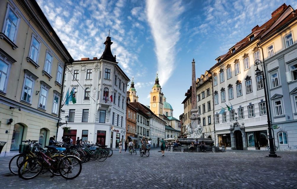 Ampia veduta di una piazza acciottolata europea, con edifici storici, una fontana, un obelisco e biciclette parcheggiate.