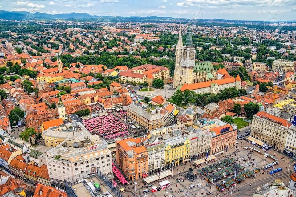 Vista aerea di una città europea con edifici dai tetti arancioni, una cattedrale a doppia guglia e una piazza del mercato con ombrelloni rossi.