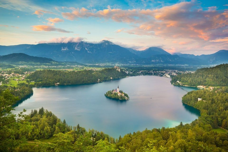 Una chiesa su una piccola isola si trova nel mezzo di un grande lago, circondata da foreste verdi e montagne sotto un cielo blu con nuvole rosa.