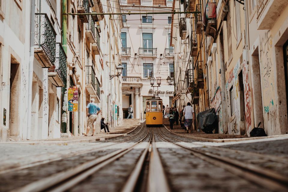 Una veduta dal basso di un tram giallo che sale una ripida e stretta strada acciottolata, fiancheggiata da edifici e pedoni.