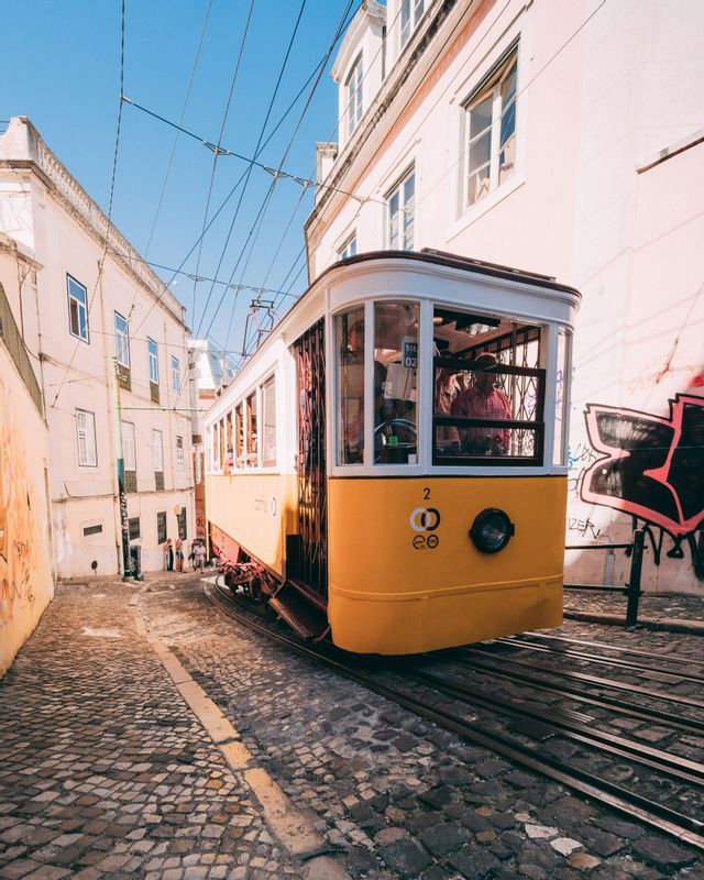Un tram giallo sale lungo una ripida e stretta strada di ciottoli, tra vecchi edifici con graffiti, sotto un cielo azzurro e limpido.