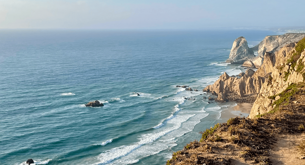 Vista dall'alto di una costa rocciosa e aspra, dove le onde blu dell'oceano si infrangono contro le scogliere e una spiaggia sabbiosa.
