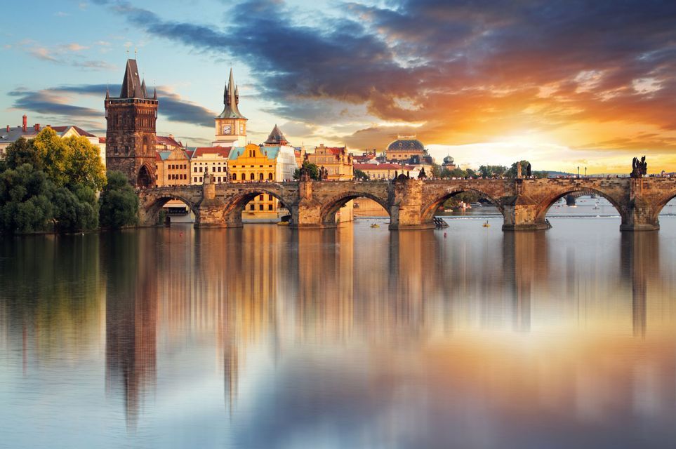 Un ponte storico in pietra e lo skyline della città adiacente si riflettono in un fiume calmo sotto un cielo colorato al tramonto.