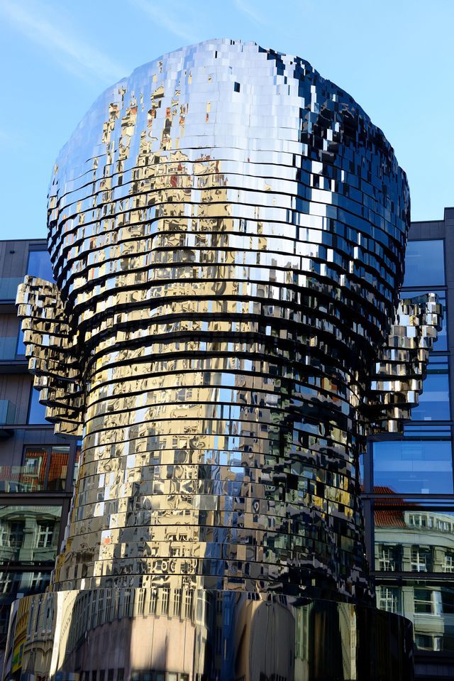 Una gran escultura cinética de una cabeza compuesta por capas apiladas y reflectantes, que reflejan los edificios circundantes y un cielo azul.