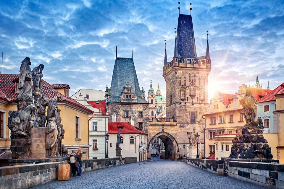 Un puente histórico de adoquines bordeado de estatuas que conduce a una puerta de torre medieval bajo un cielo nublado al amanecer.
