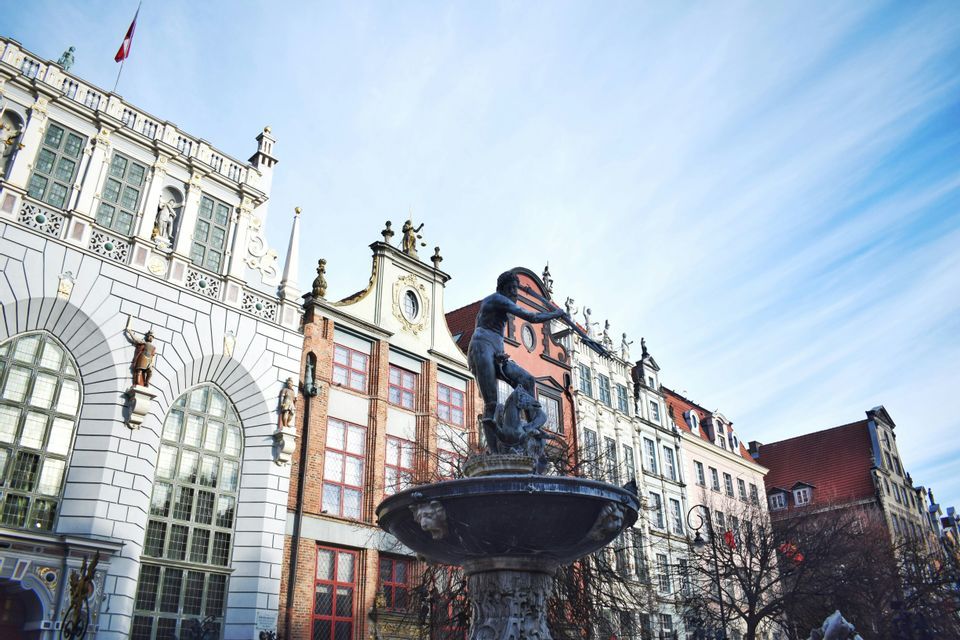 Ein bronzener Neptunbrunnen steht auf einem Stadtplatz vor verzierten, historischen Gebäuden unter blauem Himmel.