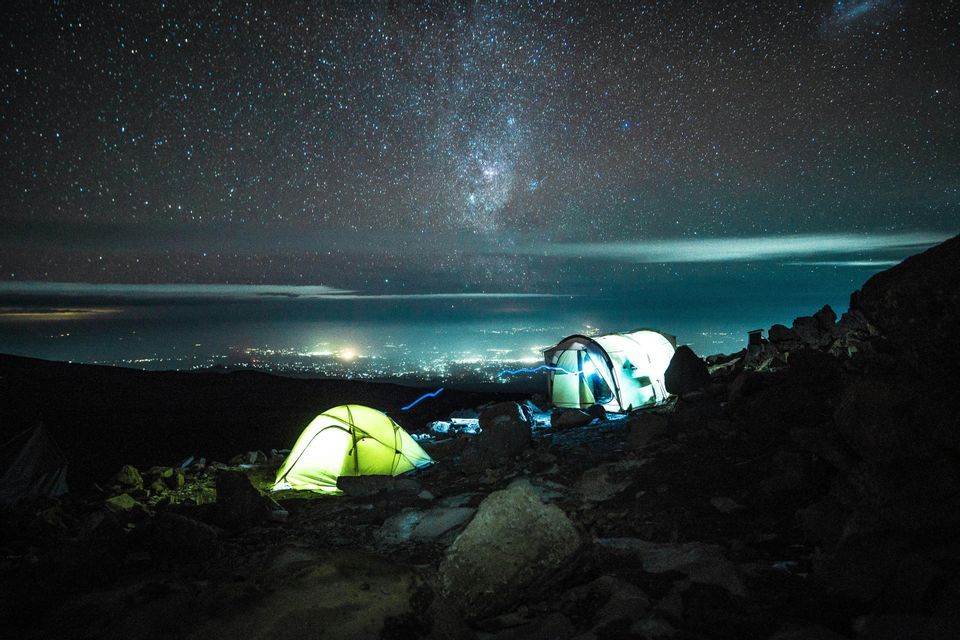 Dos tiendas iluminadas acampadas en una ladera rocosa de montaña por la noche, con la Vía Láctea visible arriba y las luces de la ciudad abajo.