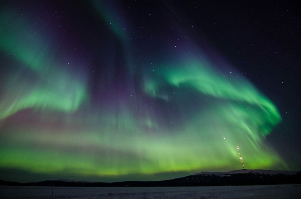 Un'aurora boreale verde e viola illumina il cielo notturno stellato sopra un paesaggio innevato con una torre distante.