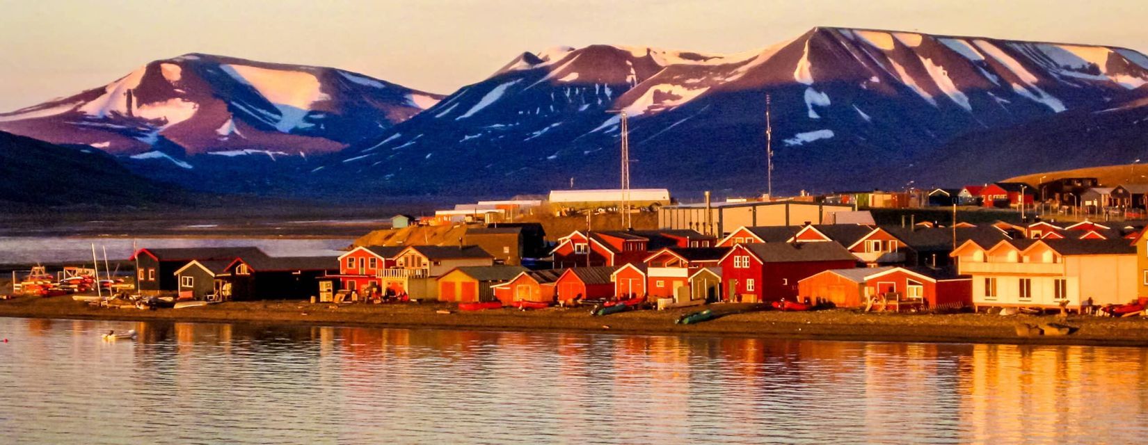 Case colorate di un villaggio di mare si riflettono nell'acqua calma, con grandi montagne innevate che si ergono sullo sfondo al tramonto.