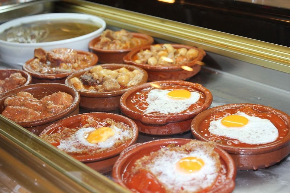 Un surtido de platos tipo tapas en cuencos de barro, algunos con huevo frito, expuestos en un mostrador de comida.