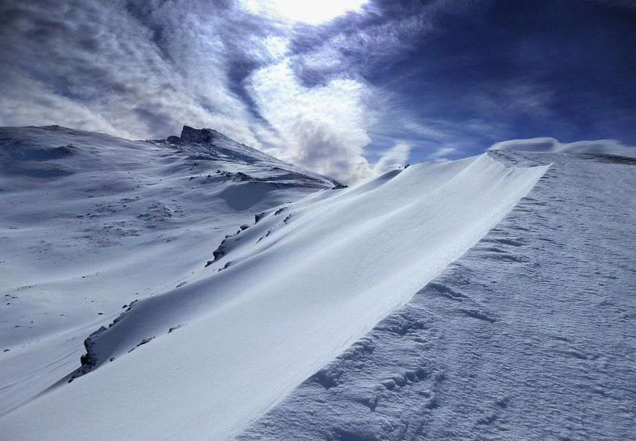 Un paisaje montañoso cubierto de nieve con una cresta barrida por el viento bajo un cielo azul profundo con nubes arremolinadas.