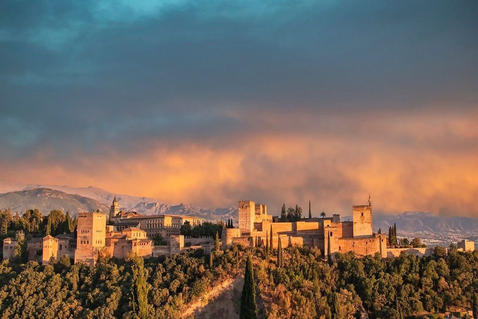 Una fortaleza de piedra histórica en una colina boscosa es iluminada por un atardecer dorado, con montañas visibles al fondo.