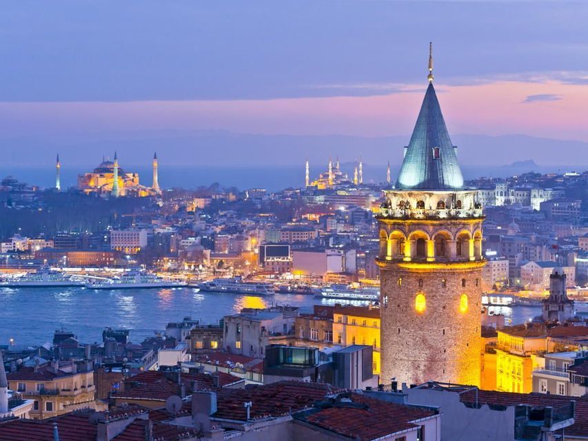 Una torre di pietra illuminata con tetto conico domina un'ampia città e il suo lungomare al tramonto.