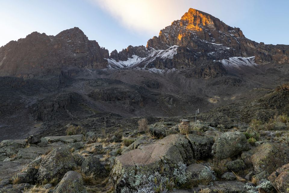 Un pico montañoso rocoso con parches de nieve, iluminado por la luz dorada del amanecer, visto desde un paisaje salpicado de rocas.