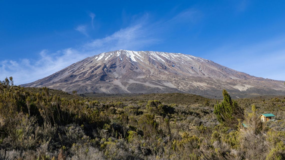 Una vista amplia de una montaña nevada que se alza sobre un paisaje de arbustos y una pequeña cabaña de madera bajo un cielo azul claro.