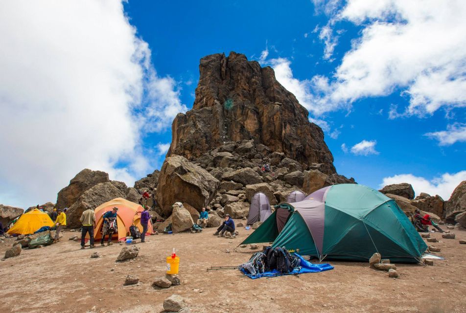 Un viaje en grupo de WeRoad acampando con tiendas de campaña en la base de una montaña empinada y rocosa bajo un cielo azul nublado.