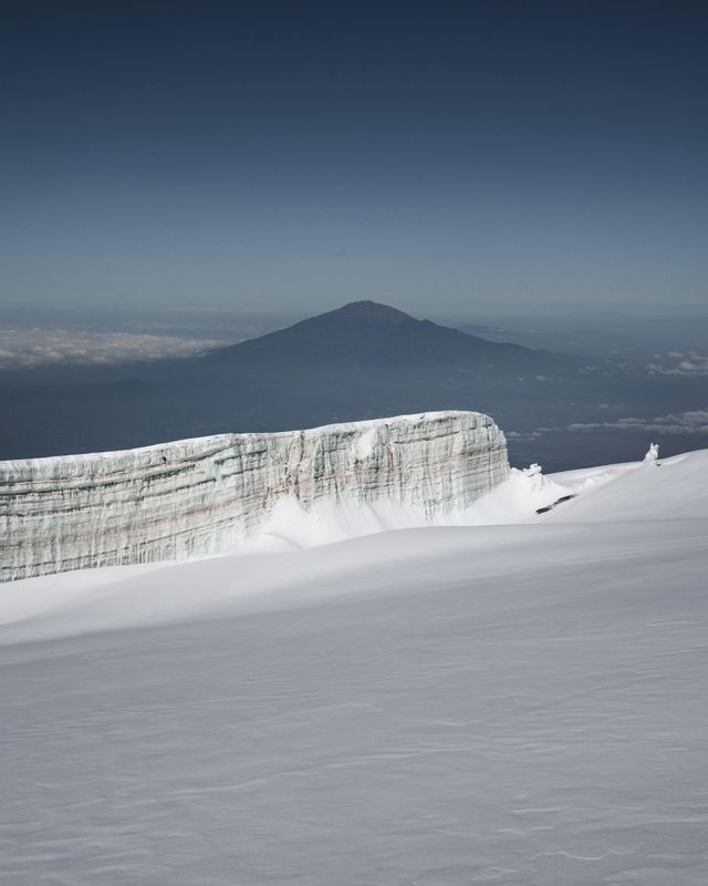 Una pared de glaciar estratificada se asienta sobre una vasta extensión cubierta de nieve, con un pico de montaña distante visible bajo un cielo despejado.
