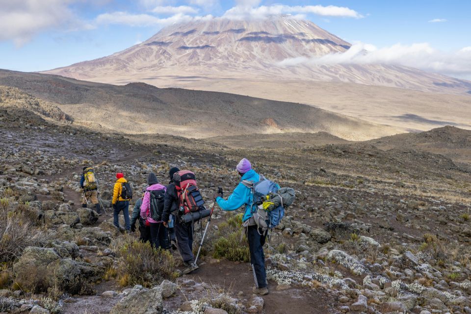 Un viaje en grupo WeRoad con mochilas, haciendo senderismo por un sendero rocoso hacia una imponente montaña a lo lejos.