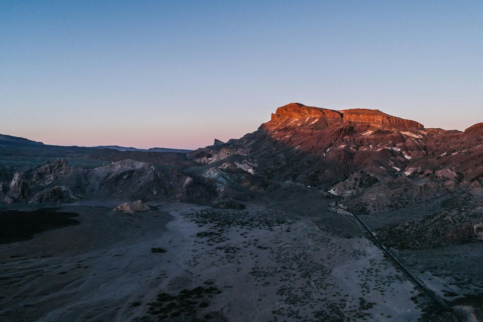 Vista aérea de un vasto paisaje árido donde un pico de montaña es iluminado por la cálida luz del amanecer bajo un cielo despejado.