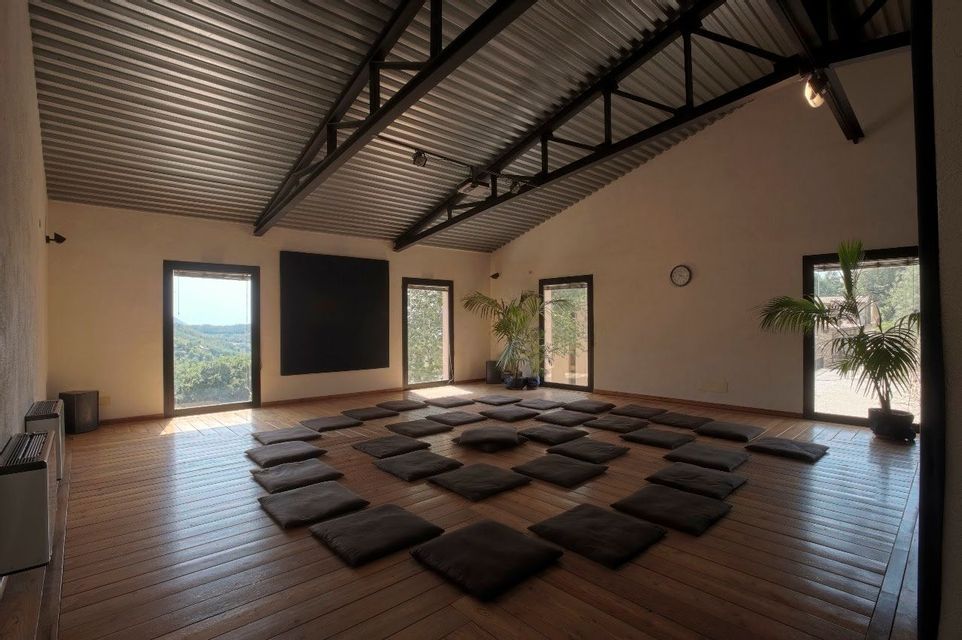 Cuscini neri disposti in cerchio sul pavimento in legno di una spaziosa e soleggiata sala di meditazione con ampie finestre.