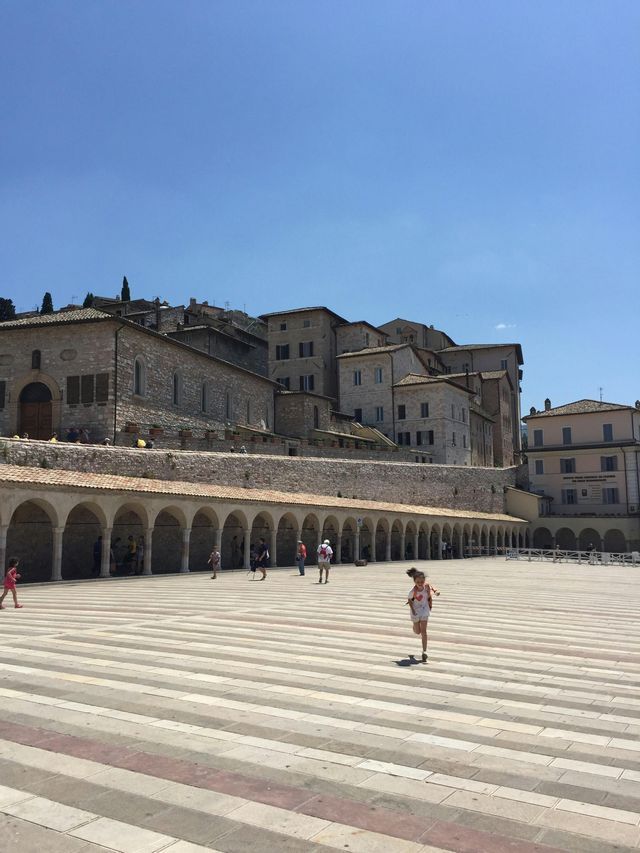 Una giovane ragazza corre su una grande piazza di pietra a strisce, di fronte a edifici storici con un colonnato ad arco, sotto un cielo soleggiato.