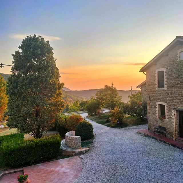Una casa di campagna in pietra con vialetto di ghiaia è circondata da alberi e dolci colline al tramonto.