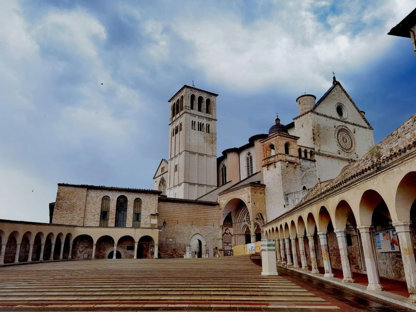 Una basilica storica con un alto campanile vista da una piazza vuota in pietra con un colonnato ad arco sotto un cielo nuvoloso.