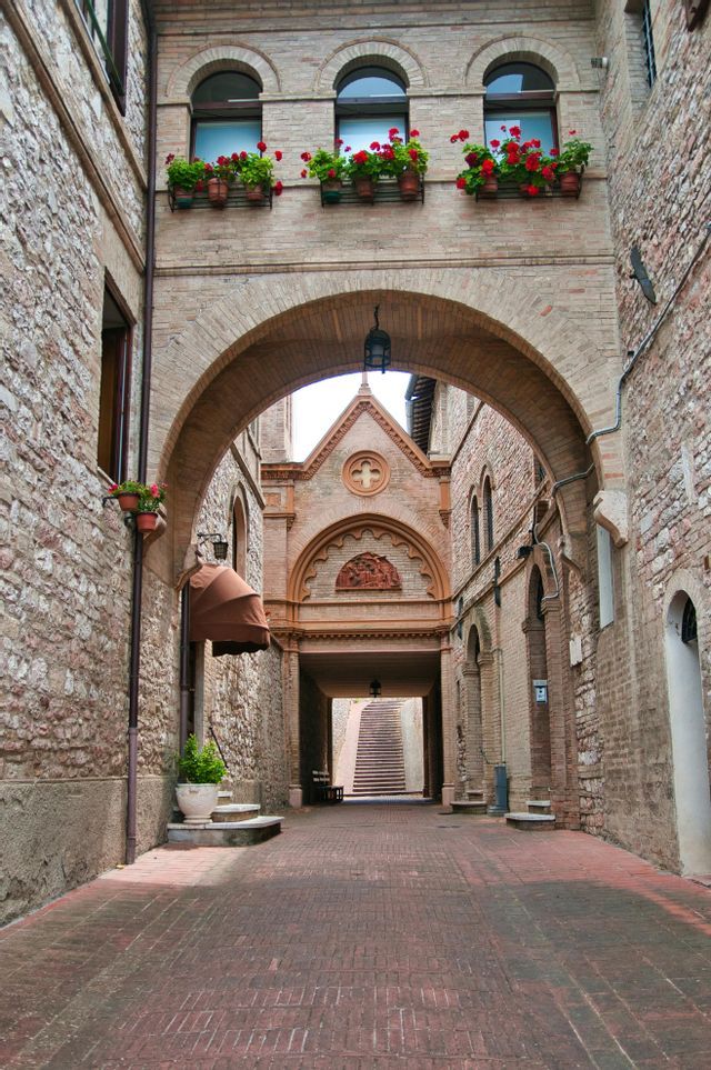 Un arco in mattoni rossi attraversa uno stretto vicolo in pietra, con fiori rossi in vaso sulle finestre soprastanti e una scala in lontananza.