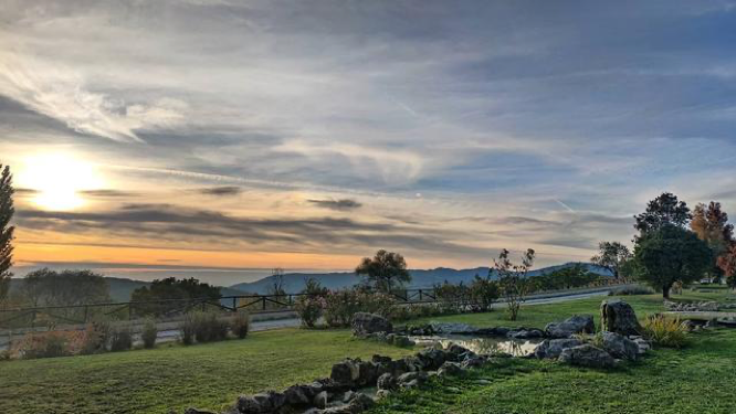 Una vista panoramica da una collina erbosa con un piccolo laghetto roccioso, che si affaccia su montagne lontane sotto un tramonto nuvoloso.
