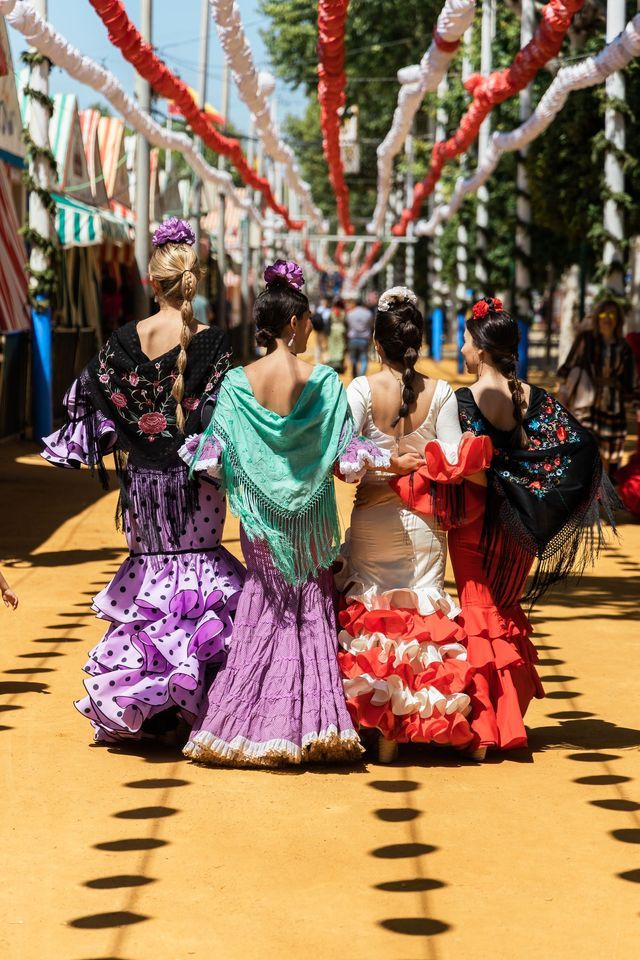 Da dietro, quattro donne in abiti da flamenco colorati e svolazzanti camminano lungo una strada gialla sotto ghirlande festive.