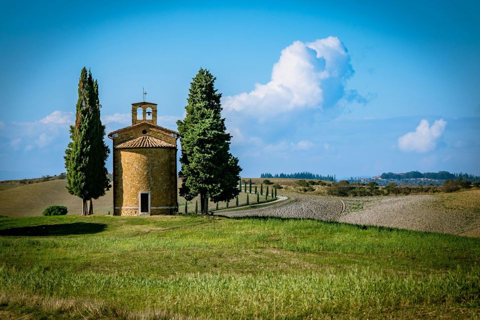 Una cappella in pietra con campanile sorge tra due alti cipressi su una collina erbosa, con campi ondulati sullo sfondo.