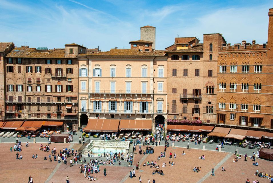 Una vista dall'alto di un'ampia piazza europea soleggiata, piena di persone che camminano e siedono vicino a una fontana centrale e a edifici storici.