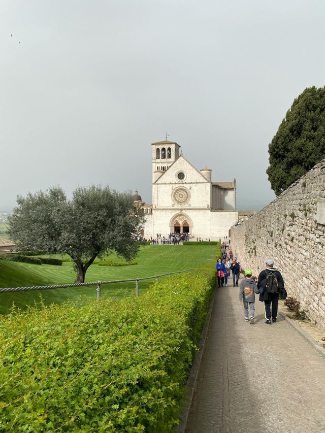 Le persone camminano lungo un sentiero di pietra verso una grande chiesa di pietra bianca con un rosone, accanto a un prato verde curato.