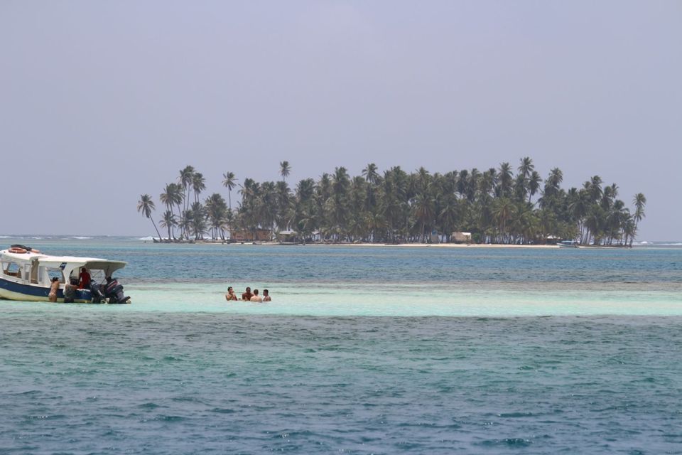 A WeRoad group trip relaxes in shallow turquoise water by a boat, with a tropical island covered in palm trees in the background.
