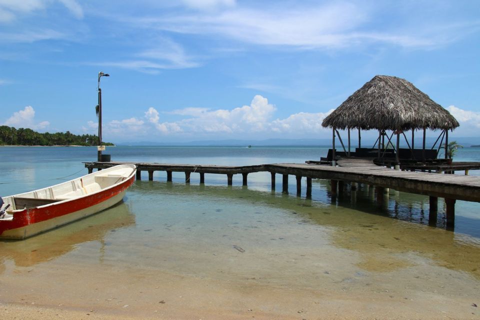 A red and white boat is moored to a wooden pier with a thatched-roof hut over clear tropical water under a blue sky.