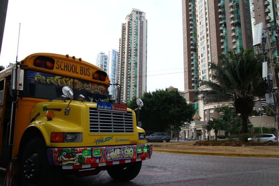 A yellow school bus with colorful graffiti art is parked on a city street, with modern high-rise buildings in the background.
