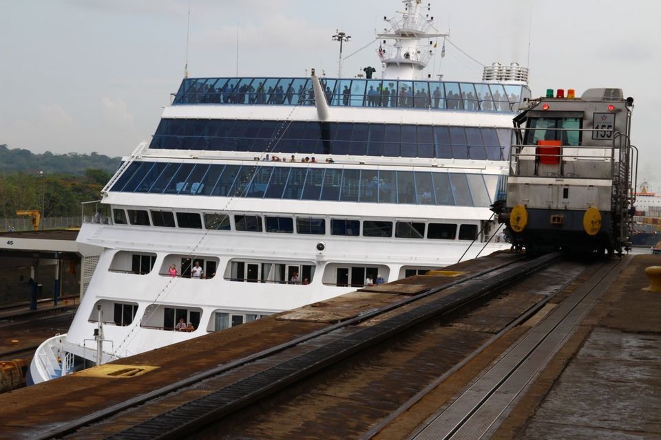 A mule train on tracks pulls a large cruise ship through a canal lock while passengers watch from the upper decks.