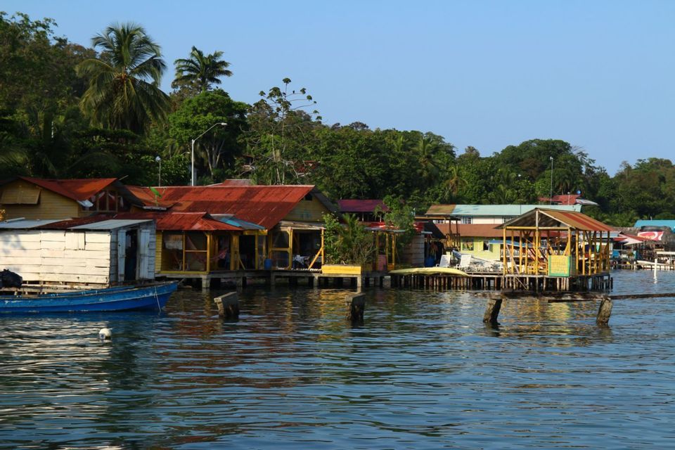 A waterfront village with colorful houses on stilts, on the edge of a lush forest, with a small blue boat moored nearby.