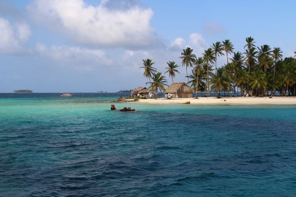 View from the water of a tropical island with huts and palm trees on a white sand beach under a cloudy sky.