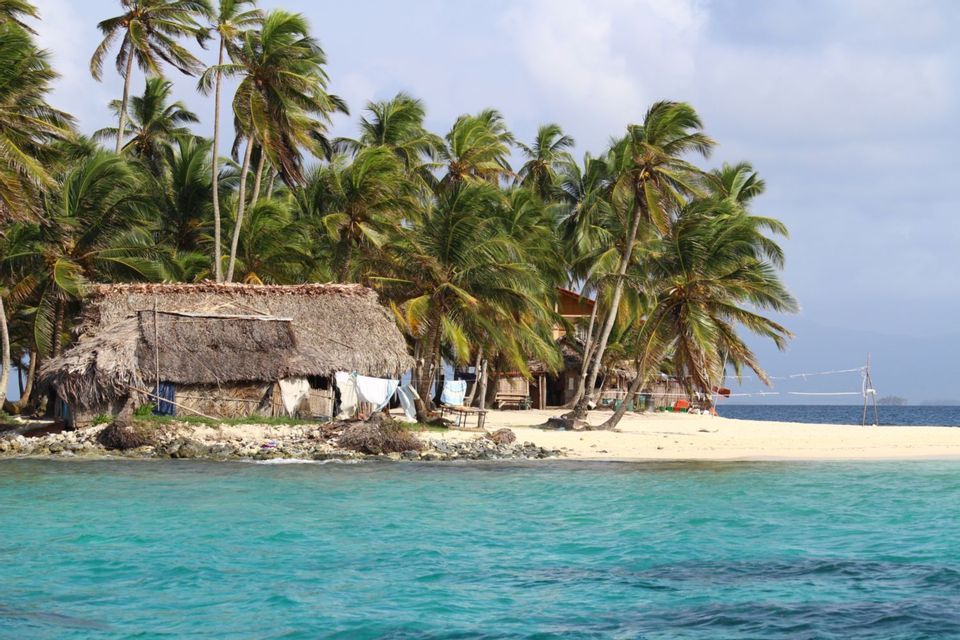A thatched-roof hut stands on a small, sandy island surrounded by palm trees and clear turquoise water.