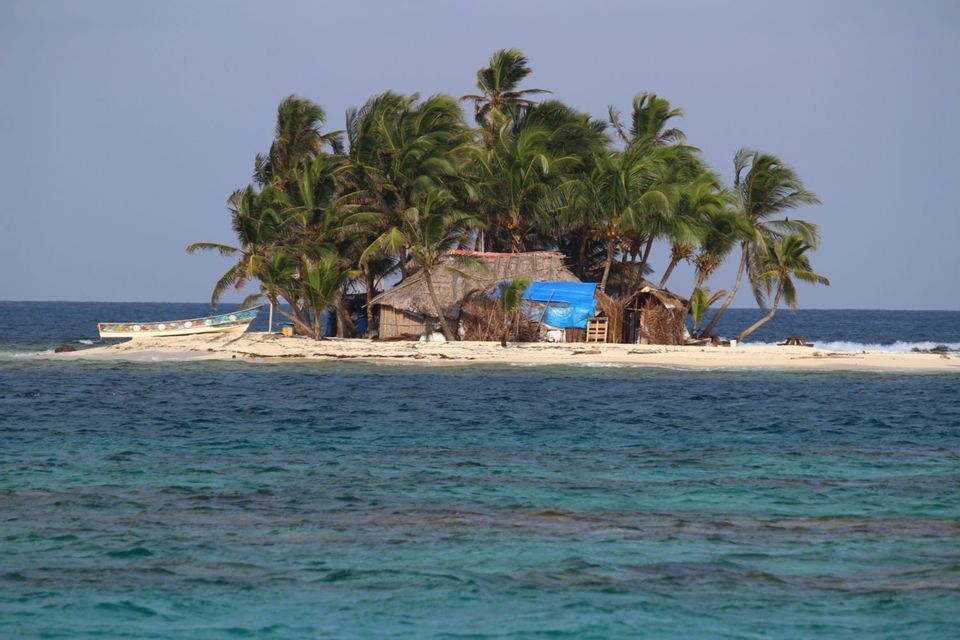 A small sandy island with palm trees, thatched huts, and a beached boat, surrounded by turquoise and deep blue ocean water.