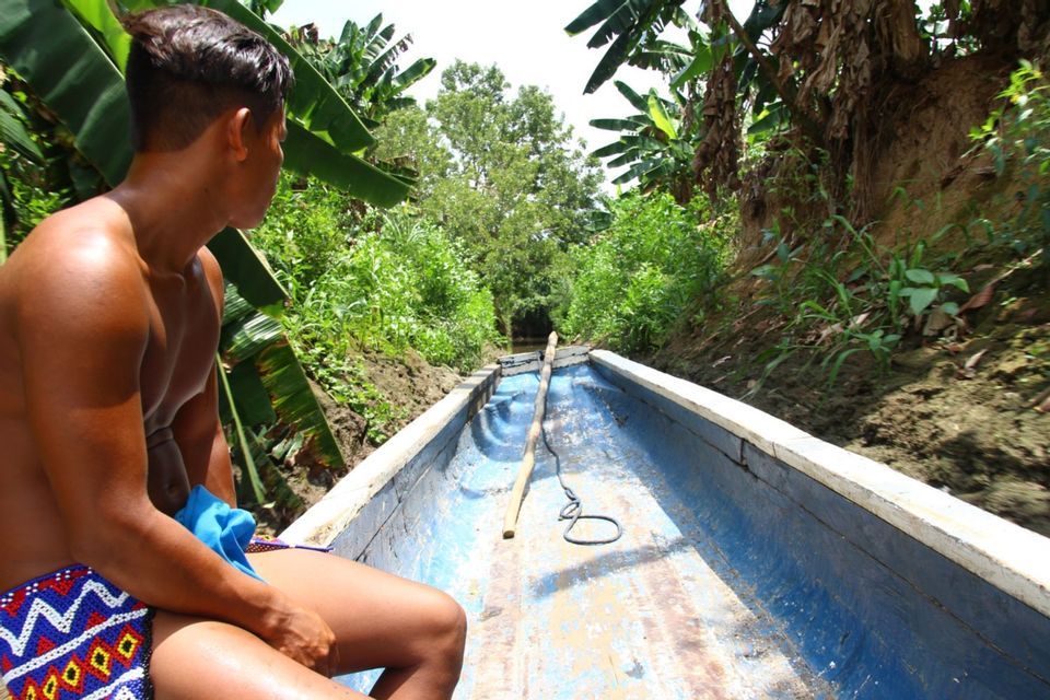 A man in patterned shorts sits in a long blue boat, sailing down a narrow canal surrounded by tropical vegetation.