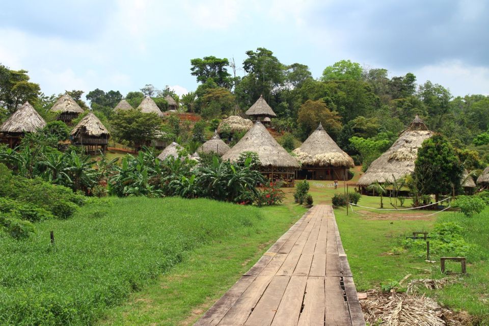 A wooden walkway leads through a grassy field toward a village of huts with thatched roofs, set on a lush, green hillside.