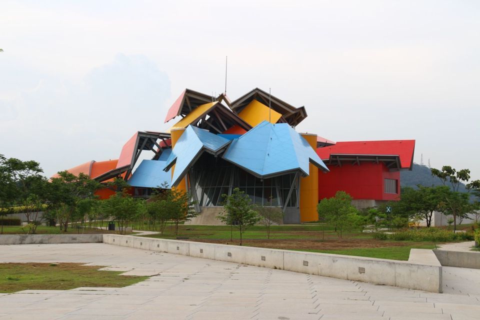 A modern building with angular roofs colored in red, blue and yellow, located in a park under a cloudy sky.