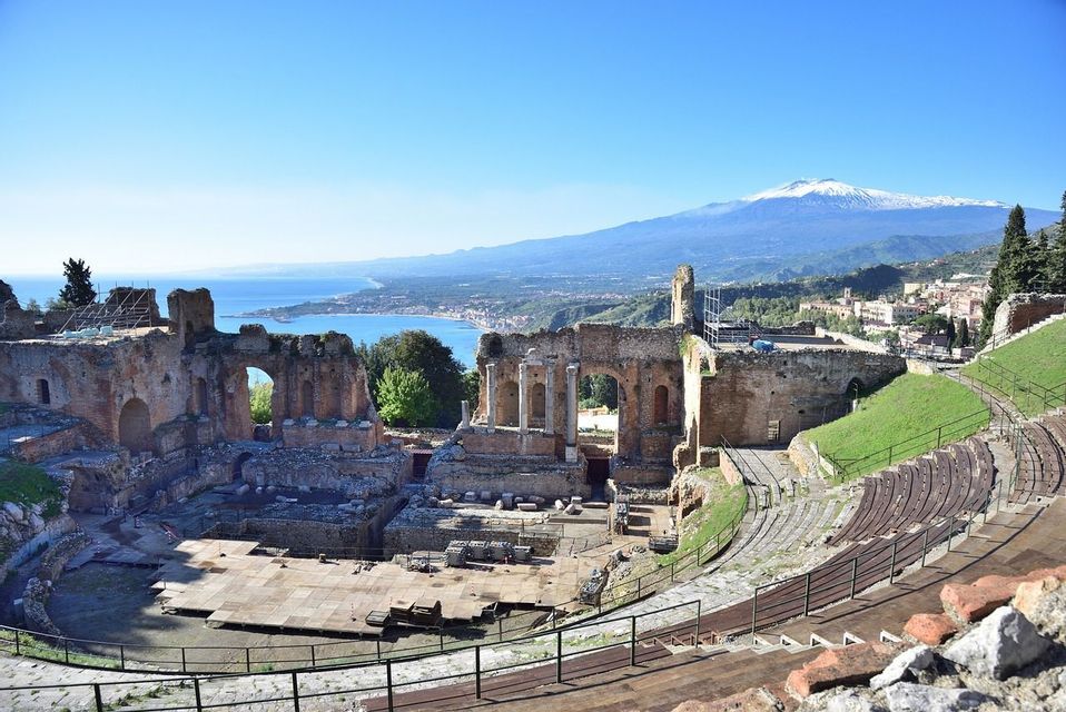 Le rovine di un antico anfiteatro con vista su una costa e una montagna innevata in lontananza, sotto un cielo azzurro e limpido.
