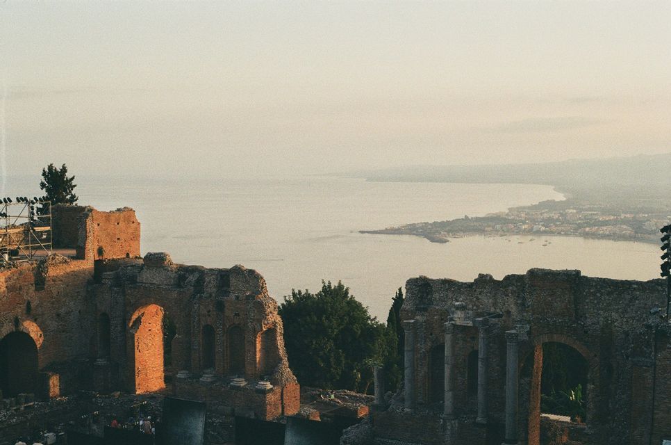 Le rovine di un anfiteatro in pietra che si affacciano su una grande baia con una città costiera al tramonto.
